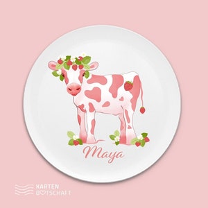 May include: A white ceramic plate with a pink and white cow wearing a strawberry crown. The cow is standing on a bed of green leaves and strawberries. The plate has the name "Maya" written in pink cursive lettering.