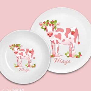 May include: Two white ceramic plates with a pink cow design. The larger plate has the name "Maya" written on it. The smaller plate has a smaller pink cow and the name "Maya" written on it.