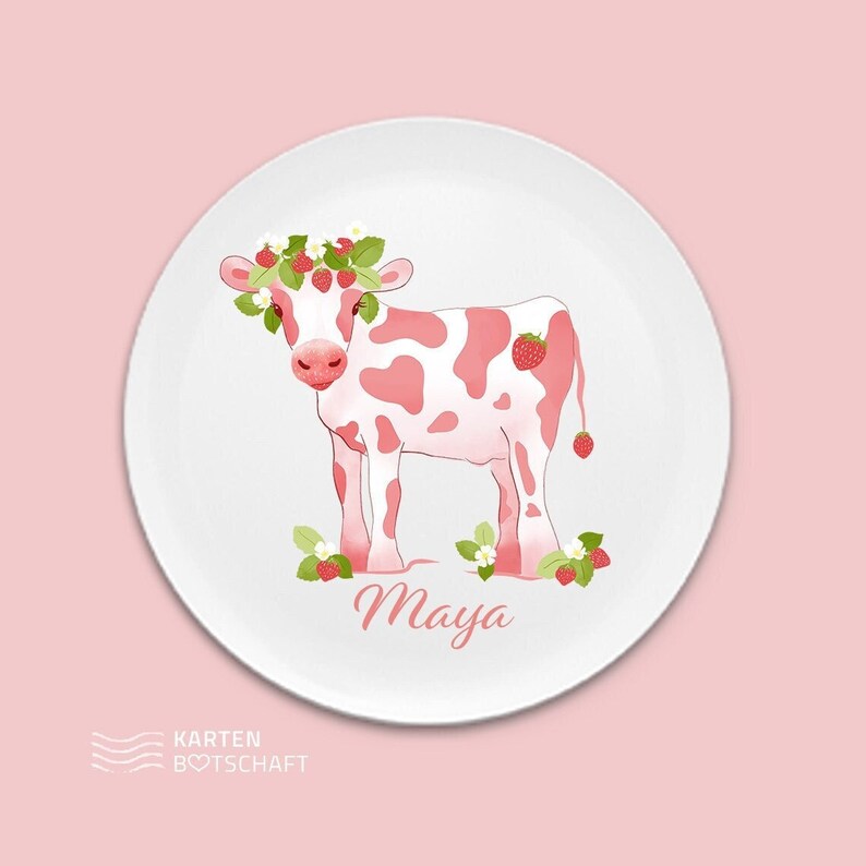 May include: A white ceramic plate with a pink and white cow wearing a strawberry crown. The cow is standing on a bed of green leaves and strawberries. The plate has the name "Maya" written in pink cursive lettering.
