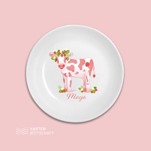 May include: A white ceramic plate with a pink cow wearing a strawberry crown. The cow is surrounded by strawberries and leaves. The plate has the name "Maya" written on it.