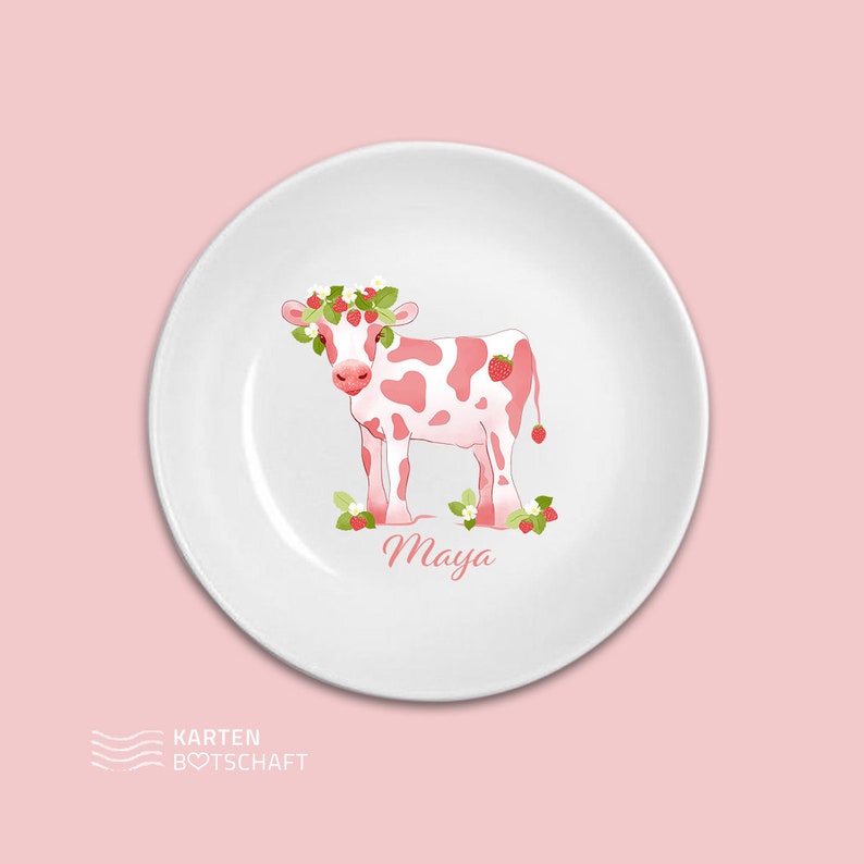 May include: A white ceramic plate with a pink cow wearing a strawberry crown. The cow is surrounded by strawberries and leaves. The plate has the name "Maya" written on it.