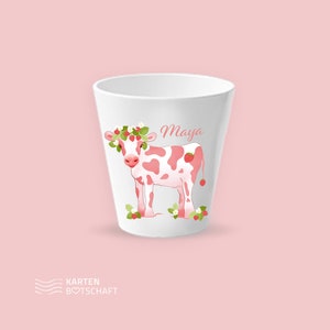 May include: A white plastic cup with a pink and white cow with a strawberry crown and the name "Maya" printed on it.