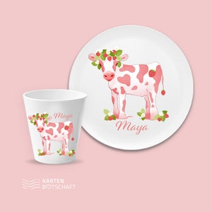 May include: A white ceramic plate and cup with a pink cow wearing a strawberry crown. The cow is surrounded by strawberries and flowers. The plate and cup both have the name "Maya" written on them.