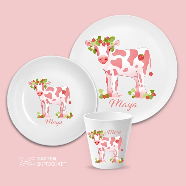 May include: A pink and white cow with a strawberry crown on a white plate. The plate has the name "Maya" written on it. There is a smaller plate and a cup with the same design.