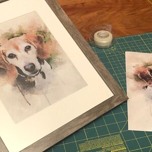 May include: A framed watercolor painting of a dog with brown and white fur. The dog is looking directly at the camera. The painting is on a white mat and has a gray wood frame.