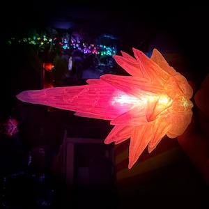 May include: A translucent, crystalline-shaped object emitting a vibrant glow that transitions from a deep magenta at the pointed tip to a warm, bright orange at the base, featuring sharp, angular facets and a textured, layered surface structure.