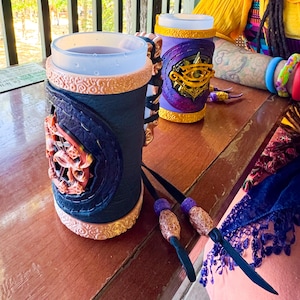 May include: Two decorative drink holders. One is navy blue with a coral and gold design, the other is purple with a gold eye symbol. Both have a clear glass insert. The holders are on a wooden surface.