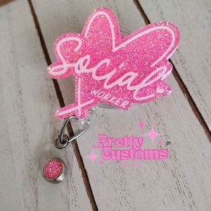 May include: A pink glittery heart-shaped badge reel featuring the white script text Social Worker. The badge reel includes a clear plastic strap with a small circular pink glitter accent and a metal clip for attachment to clothing or lanyards.