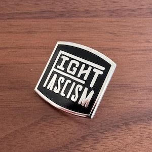Może przedstawiać: Czarna i srebrna szpilka emaliowana z napisem "Fight Fascism" białymi literami.