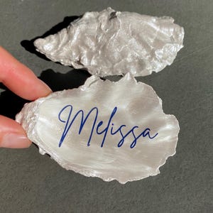 Könnte beinhalten: Zwei weiße Austernmuschel-Platzkarte mit dem Namen "Melissa" in blauer Tinte.