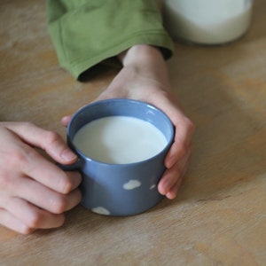 May include: A blue ceramic mug with white cloud designs filled with milk. The mug is being held by a person's hands.