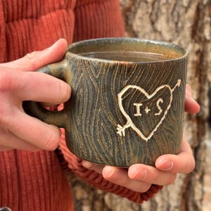May include: A handmade ceramic mug with a dark, textured finish. The mug has a carved heart design pierced by an arrow, with the initials "I + S" inside. The mug is filled with a warm drink.