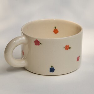 Puede incluir: Una taza de cer&aacute;mica blanca con un asa redondeada y un patr&oacute;n floral. Las flores son rosas, naranjas y azules.