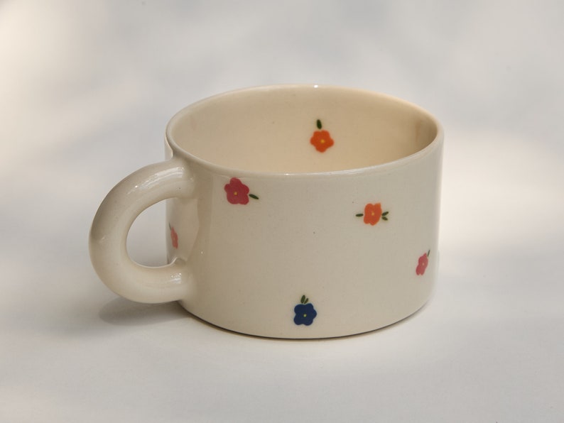 Puede incluir: Una taza de cer&aacute;mica blanca con un asa redondeada y un patr&oacute;n floral. Las flores son rosas, naranjas y azules.
