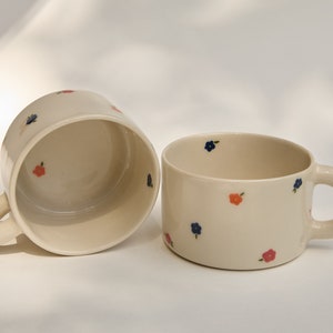 Puede incluir: Dos tazas de cer&aacute;mica blanca con peque&ntilde;os dise&ntilde;os florales azules y naranjas. Las tazas tienen asas redondeadas.