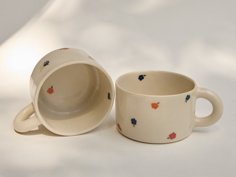 Puede incluir: Dos tazas de cer&aacute;mica blanca con peque&ntilde;os dise&ntilde;os florales azules y naranjas. Las tazas tienen asas redondeadas.