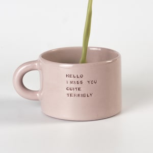 Peut inclure: Une tasse en céramique rose clair avec une anse arrondie et le texte en relief HELLO I MISS YOU QUITE TERRIBLY. Un filet de liquide vert clair est versé au centre de la tasse, remplissant le récipient sur un fond blanc uni.