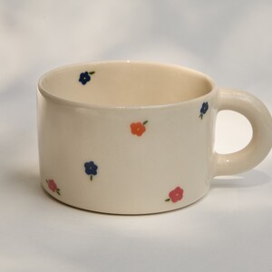 Puede incluir: Una taza de cer&aacute;mica blanca con un asa redondeada. La taza est&aacute; decorada con peque&ntilde;as flores azules y naranjas.