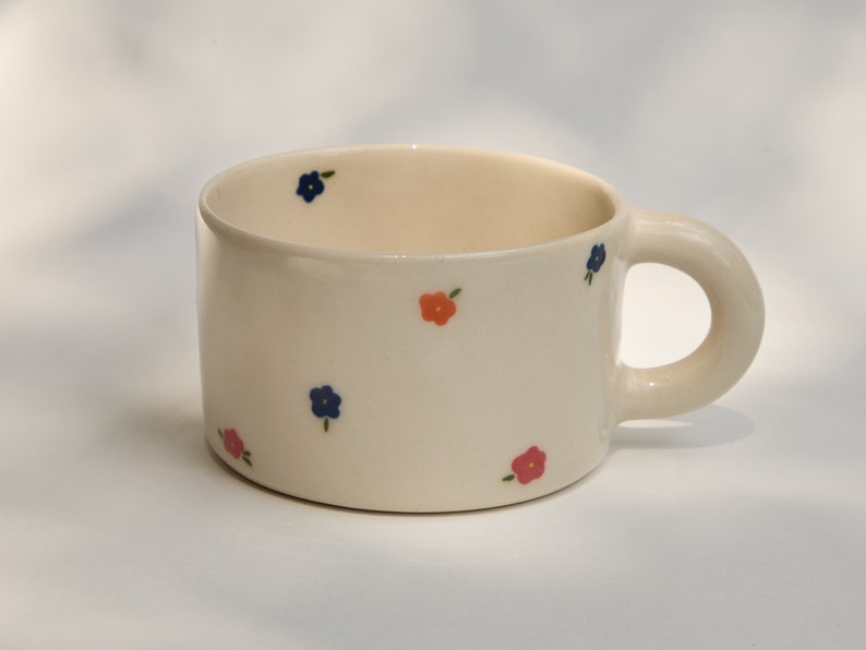 Puede incluir: Una taza de cer&aacute;mica blanca con un asa redondeada. La taza est&aacute; decorada con peque&ntilde;as flores azules y naranjas.