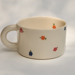 Puede incluir: Una taza de cer&aacute;mica blanca con un asa redondeada y un patr&oacute;n floral. Las flores son en tonos de rosa, naranja y azul.