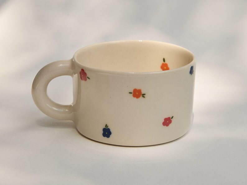 Puede incluir: Una taza de cer&aacute;mica blanca con un asa redondeada y un patr&oacute;n floral. Las flores son en tonos de rosa, naranja y azul.