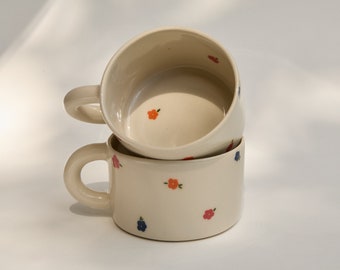 May include: Two ceramic mugs with floral designs. The mugs are stacked on top of each other. The mugs are white with small red, orange, and blue flowers.