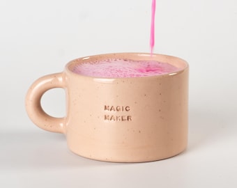 May include: A light peach-colored ceramic mug with a speckled finish and a rounded handle. The text MAGIC MAKER is embossed on the front. A bright pink liquid is being poured into the mug, creating a layer of foam on the surface of the drink.