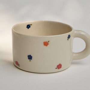 Puede incluir: Una taza de cer&aacute;mica blanca con asa, decorada con peque&ntilde;as flores azules y naranjas.