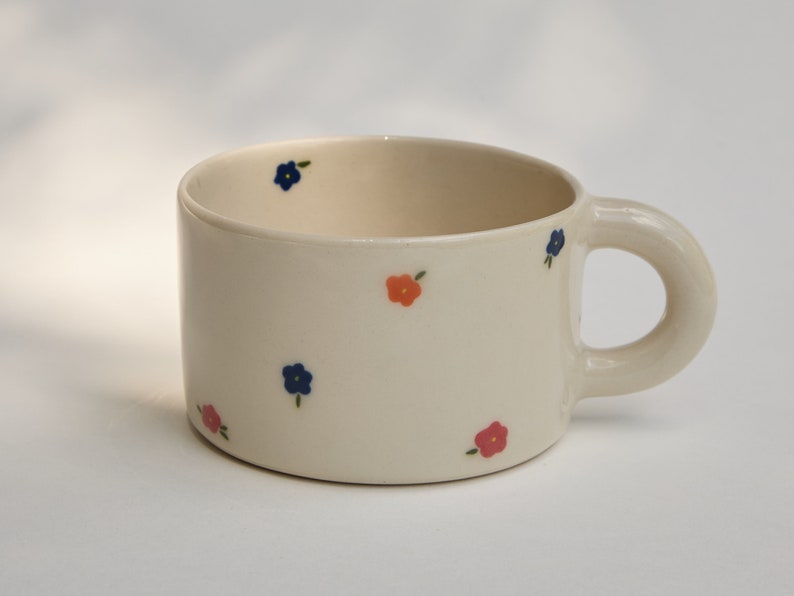 Puede incluir: Una taza de cer&aacute;mica blanca con asa, decorada con peque&ntilde;as flores azules y naranjas.