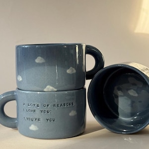 May include: Three blue ceramic mugs with white cloud designs. The bottom mug has the text "A lot's of reasons I love you: 1. You're you".