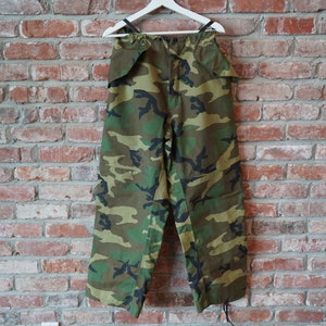 May include: Camouflage trousers hanging on a white hanger against a brick wall. The trousers feature a green, brown, and black camouflage pattern. The trousers are made of a durable material and have a relaxed fit.