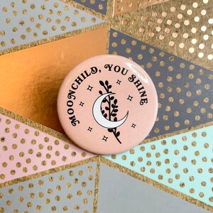 May include: A pink button with a crescent moon and a sprig of leaves. The text "MOONCHILD, YOU SHINE" is written around the moon.