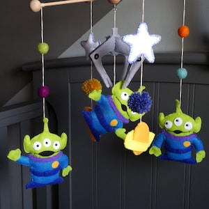 May include: A baby mobile with a space theme. It features three green aliens with three eyes each, a white star, a gray spaceship, and yellow and purple pom-poms.