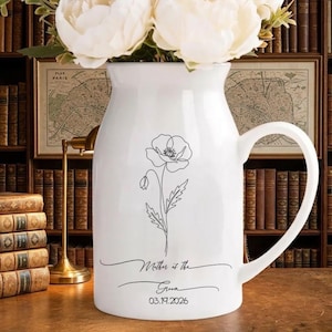 May include: White ceramic pitcher with a black floral design and the text "Mother of the Groom 03.19.2026". The pitcher has a large handle and is filled with white flowers. The background includes books and a map.