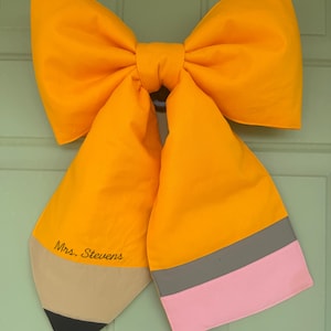 May include: A large fabric bow shaped like a pencil, featuring a bright yellow body, a tan tip with a black point, and a grey and pink eraser end. The text "Mrs. Stevens" is embroidered in black script on the yellow section of the pencil bow.