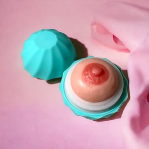 May include: A light pink and peach-colored breast-shaped object with a red nipple, set in a teal-colored container. The container is open, revealing the breast form. Another teal-colored lid is nearby. The background is pink with a pink fabric.