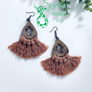 May include: A pair of brown wooden teardrop-shaped earrings with a cross cutout and a brown tassel fringe.