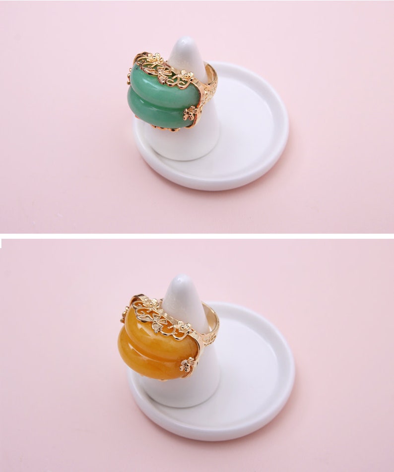 K&ouml;nnte beinhalten: Zwei Ringe mit verzierten Goldfassungen und gr&uuml;nen und gelben Edelsteineinlagen. Die Ringe werden auf wei&szlig;en Ringhaltern vor einem rosa Hintergrund pr&auml;sentiert.