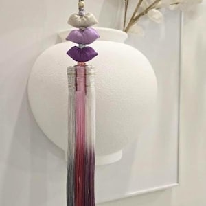 Könnte beinhalten: Ein dekorativer Quastenanhänger mit einer grauen Kordel, der mit Stoffakzenten in Beige, Lavendel und Lila geschichtet ist. Die Stränge der Quaste gehen von Rosa zu tiefem Rot und Grau über. Der Hintergrund umfasst eine weiße Vase und ein Blumenarrangement.