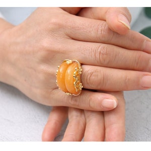 K&ouml;nnte beinhalten: Ein goldener Ring mit einem gro&szlig;en, ovalen, orangefarbenen Edelstein. Der Ring ist mit aufwendigen goldenen Details um den Stein herum verziert.