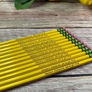 May include: A stack of yellow pencils with green and pink erasers. Each pencil is engraved with the text "Geedy's champions...we've got this!"