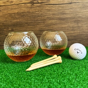 May include: Two clear glass golf ball shot glasses filled with amber liquid, sitting on a green artificial grass surface with three wooden golf tees and a white golf ball with the word "Callaway" printed on it.