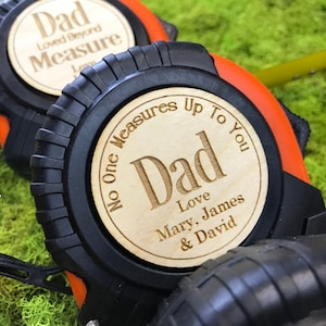 May include: Two black and orange tape measures with wooden circular accents. The wooden accents are engraved with the word "Dad" and personalized messages. One reads "Loved Beyond Measure" and the other "No One Measures Up To You".