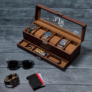 May include: A brown leather watch box with a glass top, holding several watches. The box also contains a ring, a lighter, and a beaded bracelet. Sunglasses and a watch are placed next to the box. The initials "MLC" are visible on the glass.