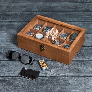 May include: A wooden watch box with a glass top, holding several wristwatches. The box is next to black sunglasses, a checkered lighter, and a carbon fiber wallet. The box has a personalized design with the name "Riley".