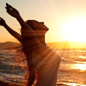 May include: Silhouette of a person with long hair standing on a beach with their arms raised in the air, facing the setting sun. The sun is shining brightly and the water is sparkling.