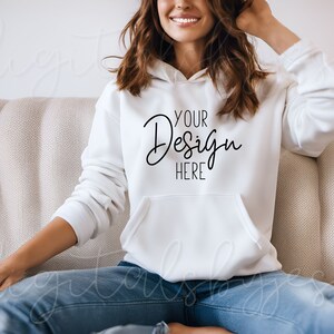 May include: A white hoodie with a large black text overlay that says "YOUR Design HERE". The hoodie is being worn by a person sitting on a couch.