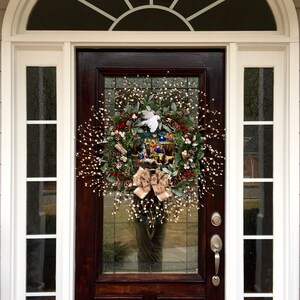 May include: A brown wooden front door with a white wreath decorated with a nativity scene and a white dove. The wreath is hanging on the door with a silver door handle and a silver deadbolt lock.