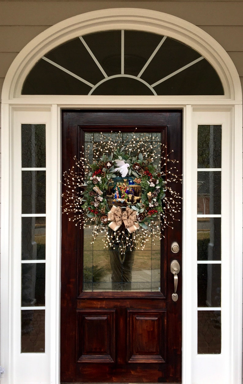May include: A brown wooden front door with a white wreath decorated with a nativity scene and a white dove. The wreath is hanging on the door with a silver door handle and a silver deadbolt lock.