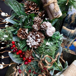 May include: A wintery wreath featuring pine branches, pinecones, red berries, and faux snow. The wreath is decorated with burlap ribbon and a pair of cinnamon sticks tied with twine.